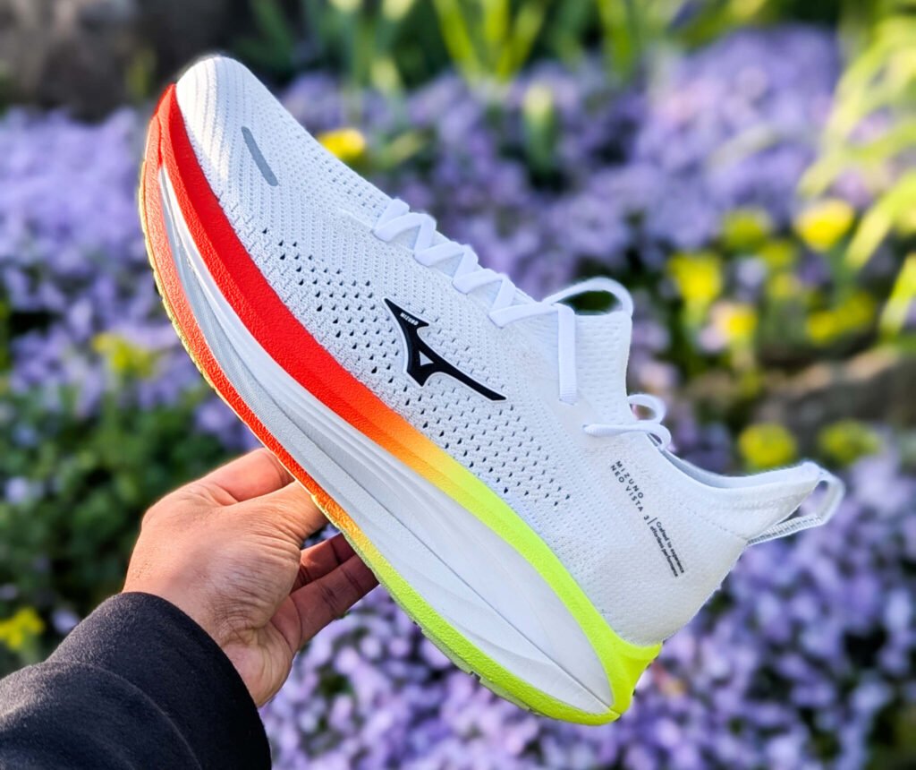 White Mizuno neo vista 3 running shoe with a bright rainbow sole, held by a hand outdoors against a blurred purple-flower background.