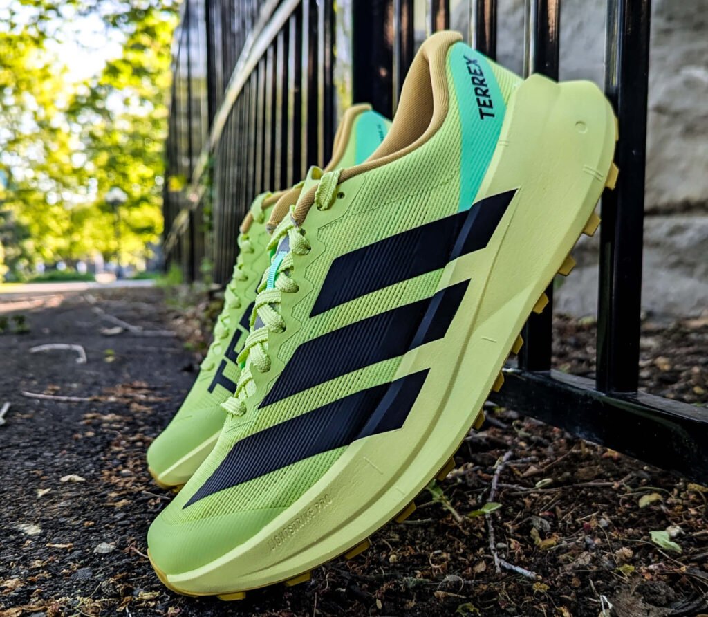 Neon green Adidas Terrex running shoes hung on a black metal fence outdoors, laces undone, sunlight filtering through trees in the background.
