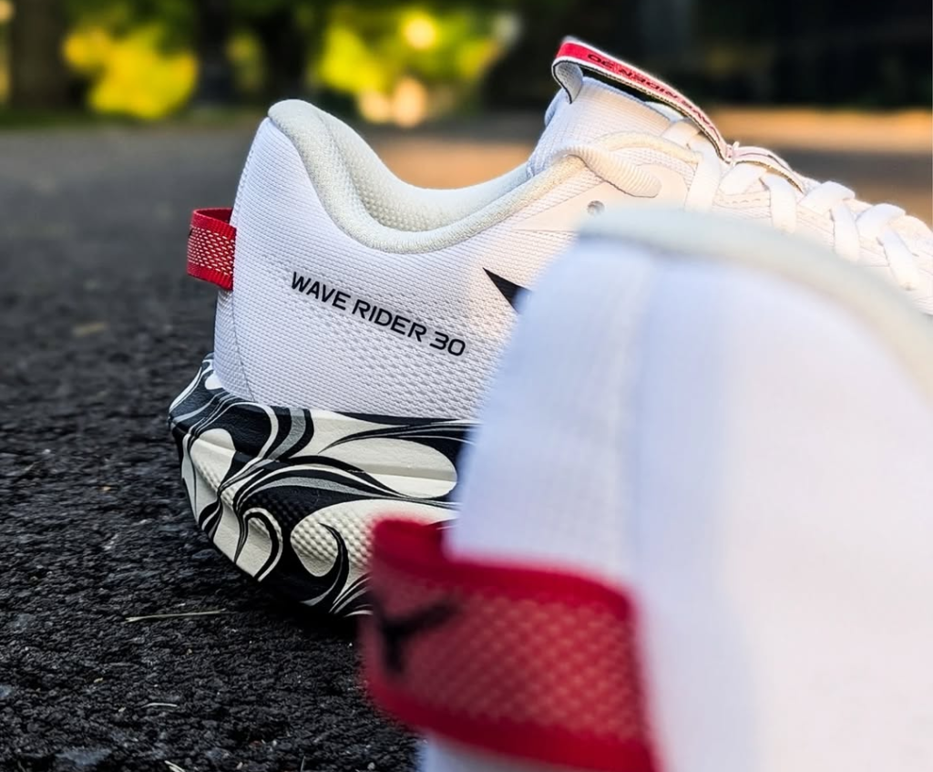 Close-up of a white Wave Rider 30 running shoe with a red heel tab resting on dark pavement; blurred green background offers outdoor lighting.